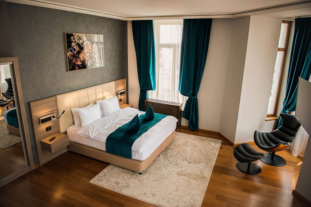 A modern bedroom with a large bed, bedside tables, a nightstand, a chair, a window with curtains, and a painting on the wall.