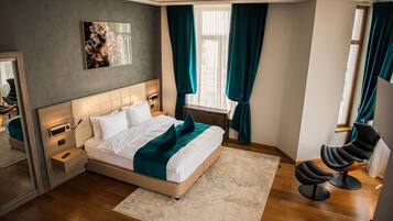 A modern bedroom with a large bed, bedside tables, a nightstand, a chair, a window with curtains, and a painting on the wall.