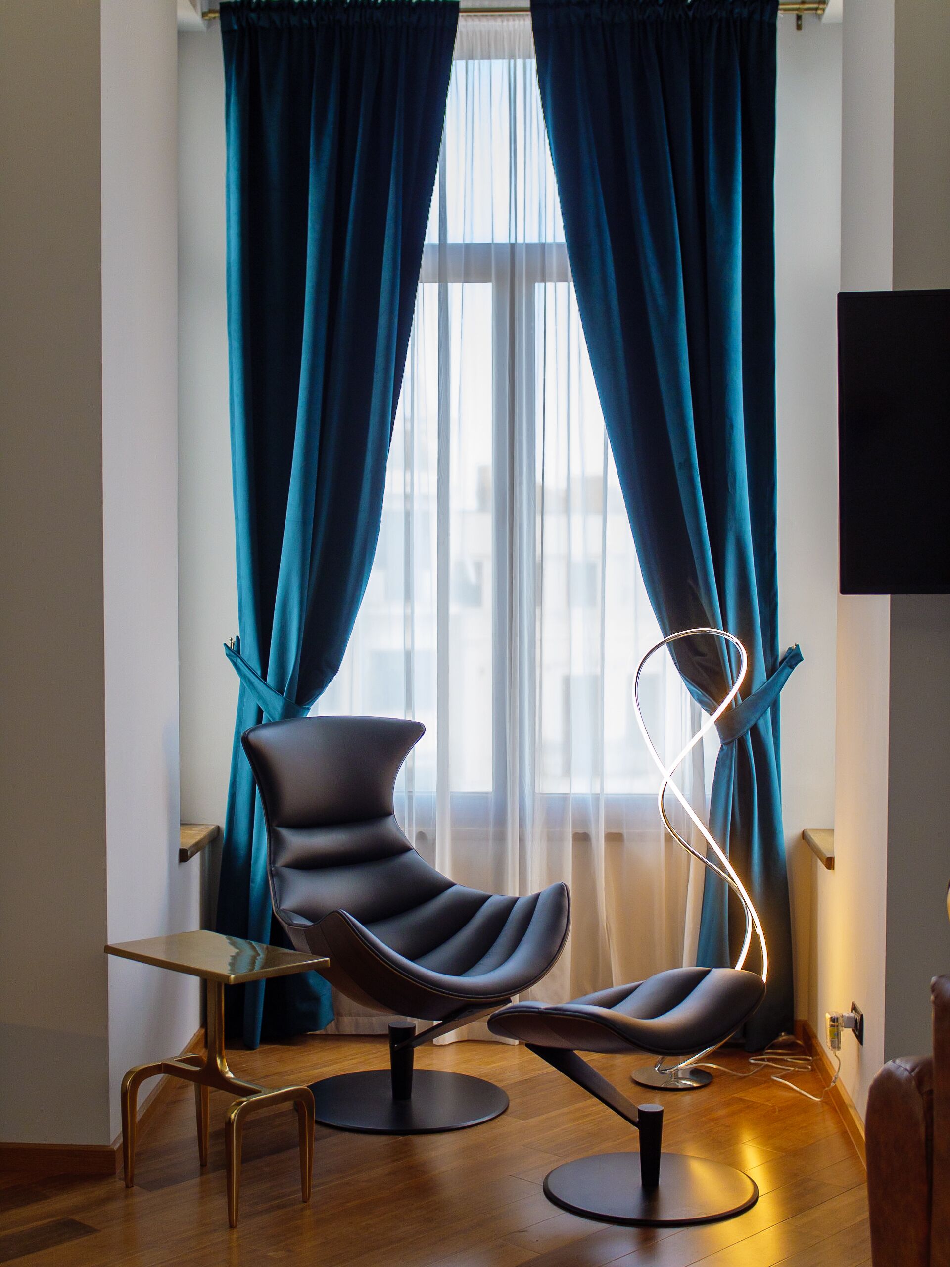 Una habitación moderna con un ventanal grande, cortinas color verde azulado, una silla negra, una mesita y una lámpara original.