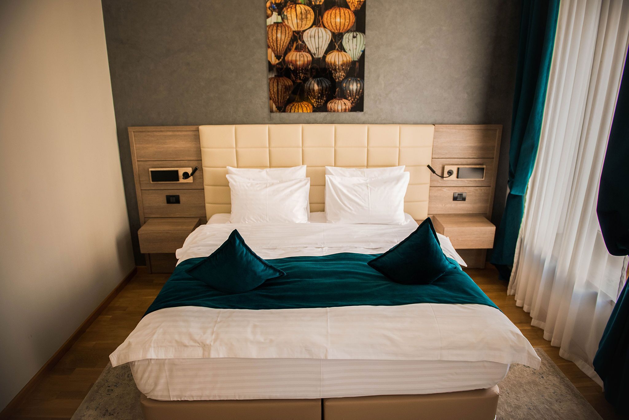 A neatly made bed with white linens and teal pillows, a wooden headboard, and a nightstand with a lamp.