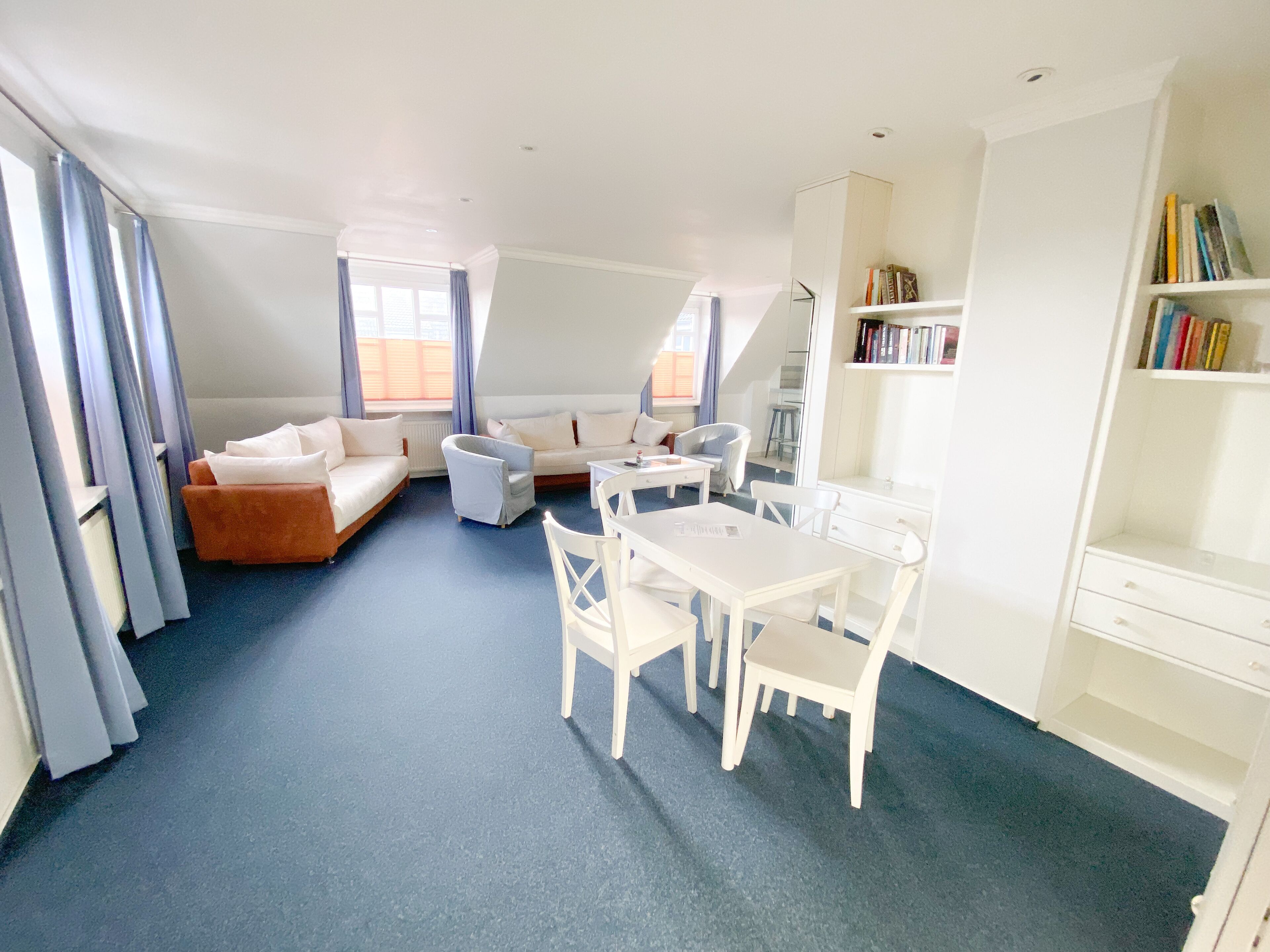 A modern, well-lit room with a white sofa set, a dining table with chairs, and a bookshelf.