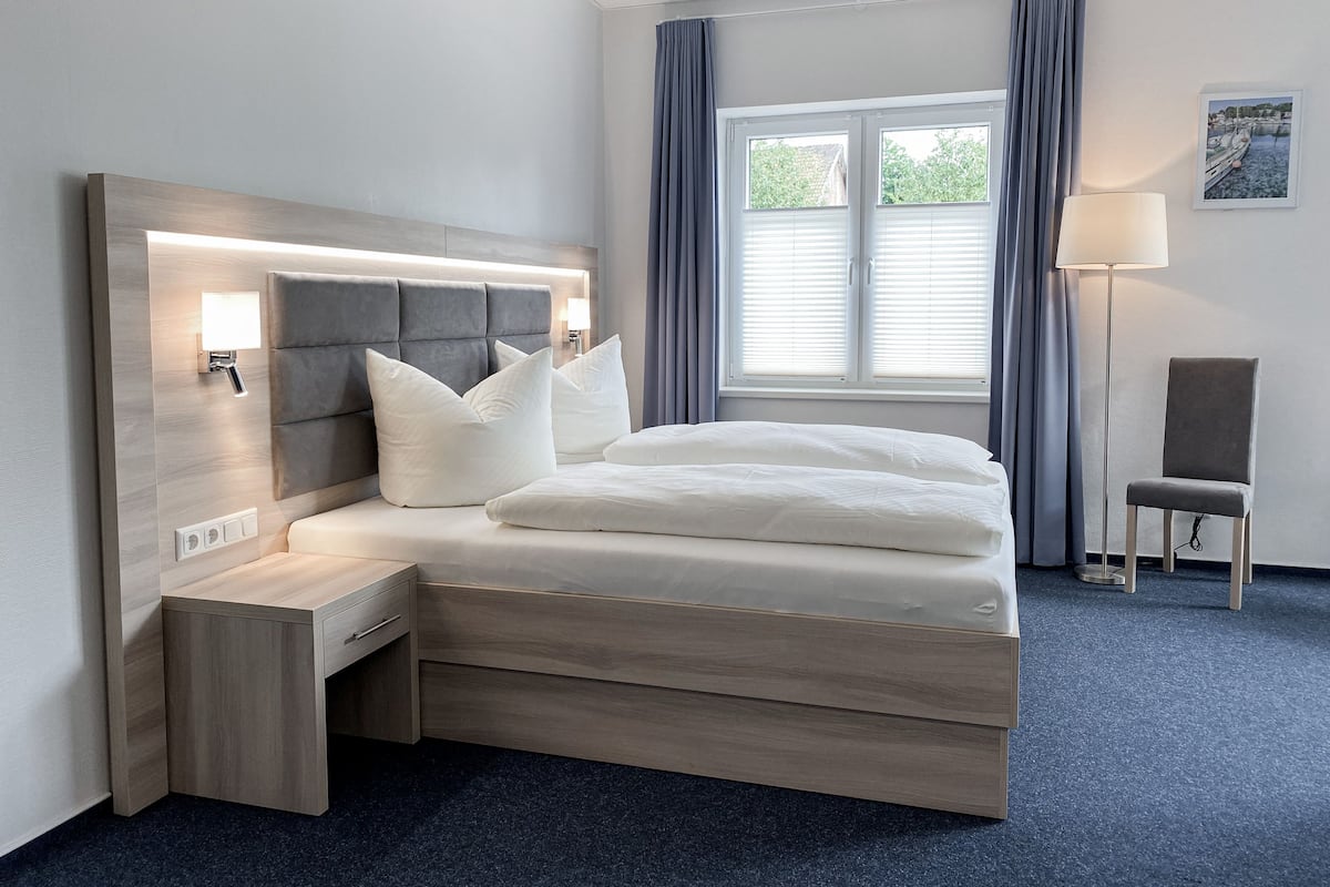 A modern hotel room with a large bed, bedside tables, a desk, a chair, a window with curtains, and a lamp.