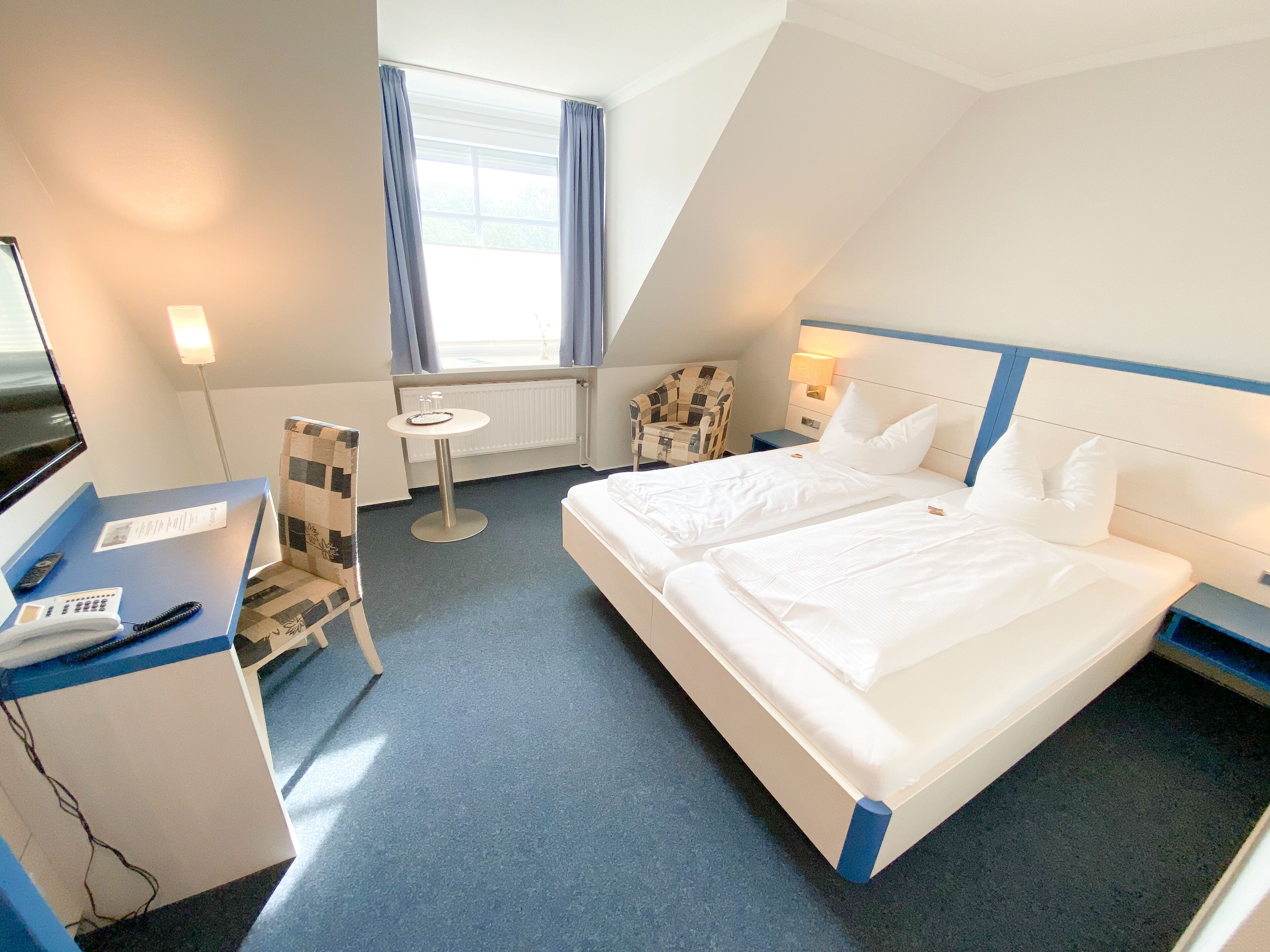 A hotel room with a bed, a desk with a telephone, a chair, a small table, and a window with curtains.