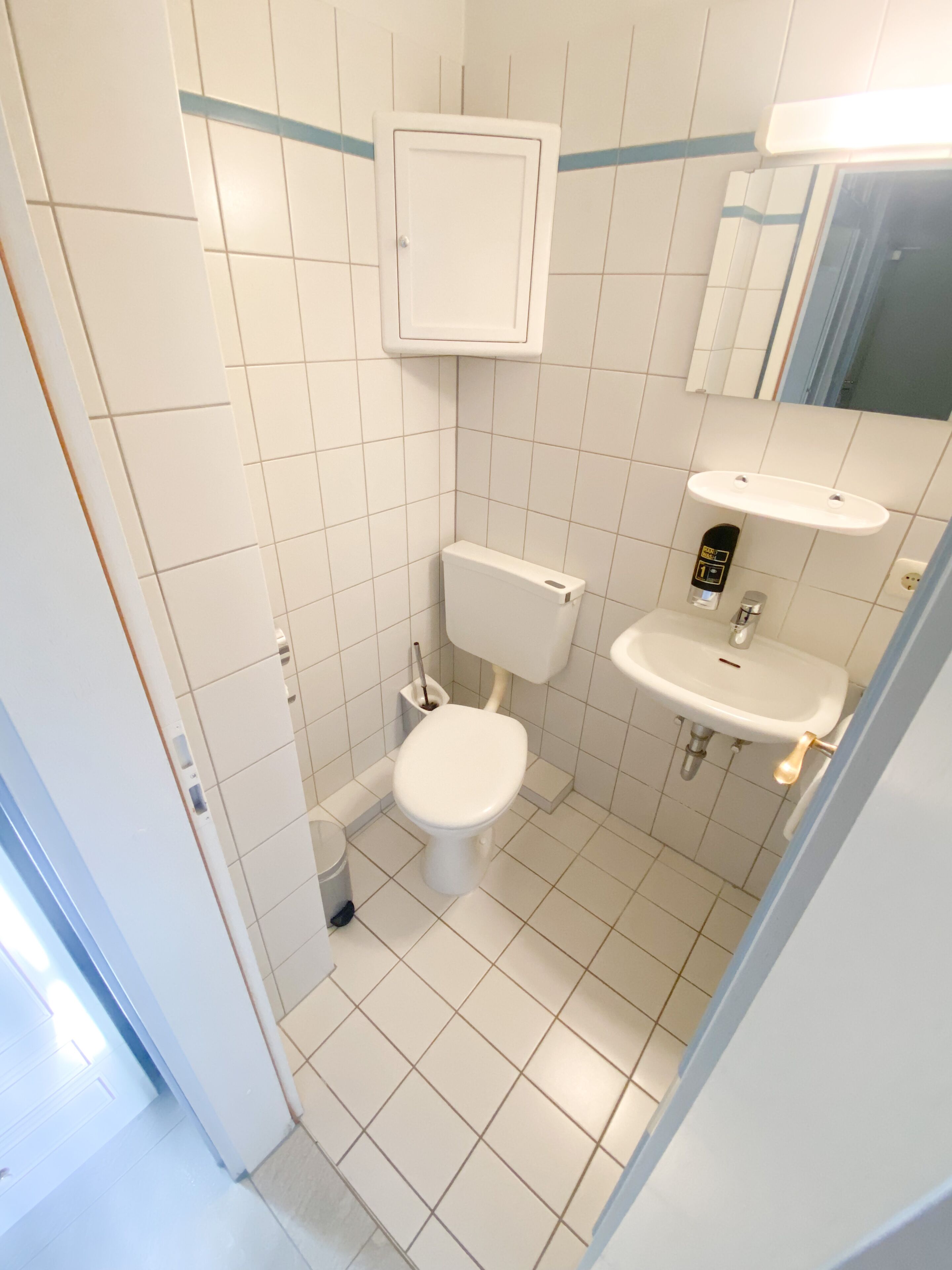 A small, clean bathroom with white tiles, a toilet, a sink, and a mirror.