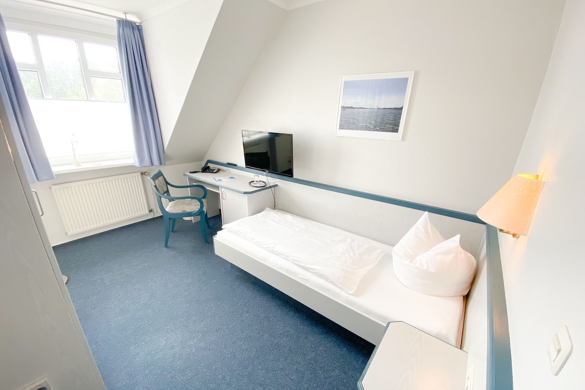 A hotel room with a single bed, a desk with a chair, a television, and a window with curtains.