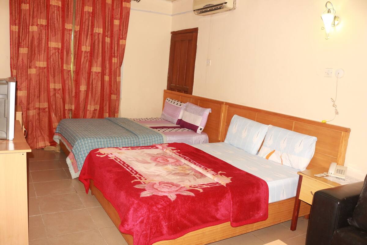 A hotel room with two beds, a desk, a chair, a TV, and an air conditioning unit.