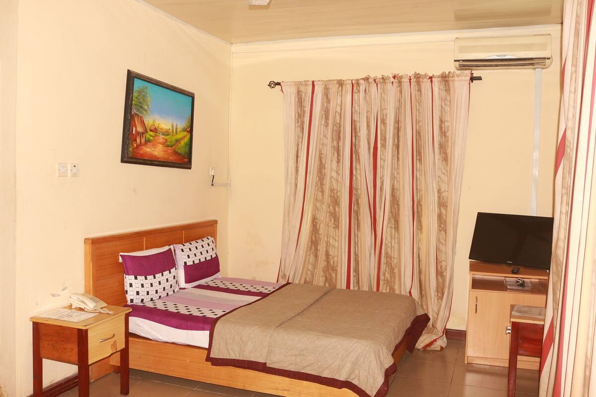 A bedroom with a bed, a bedside table with a phone, a TV on a stand, and a painting on the wall.
