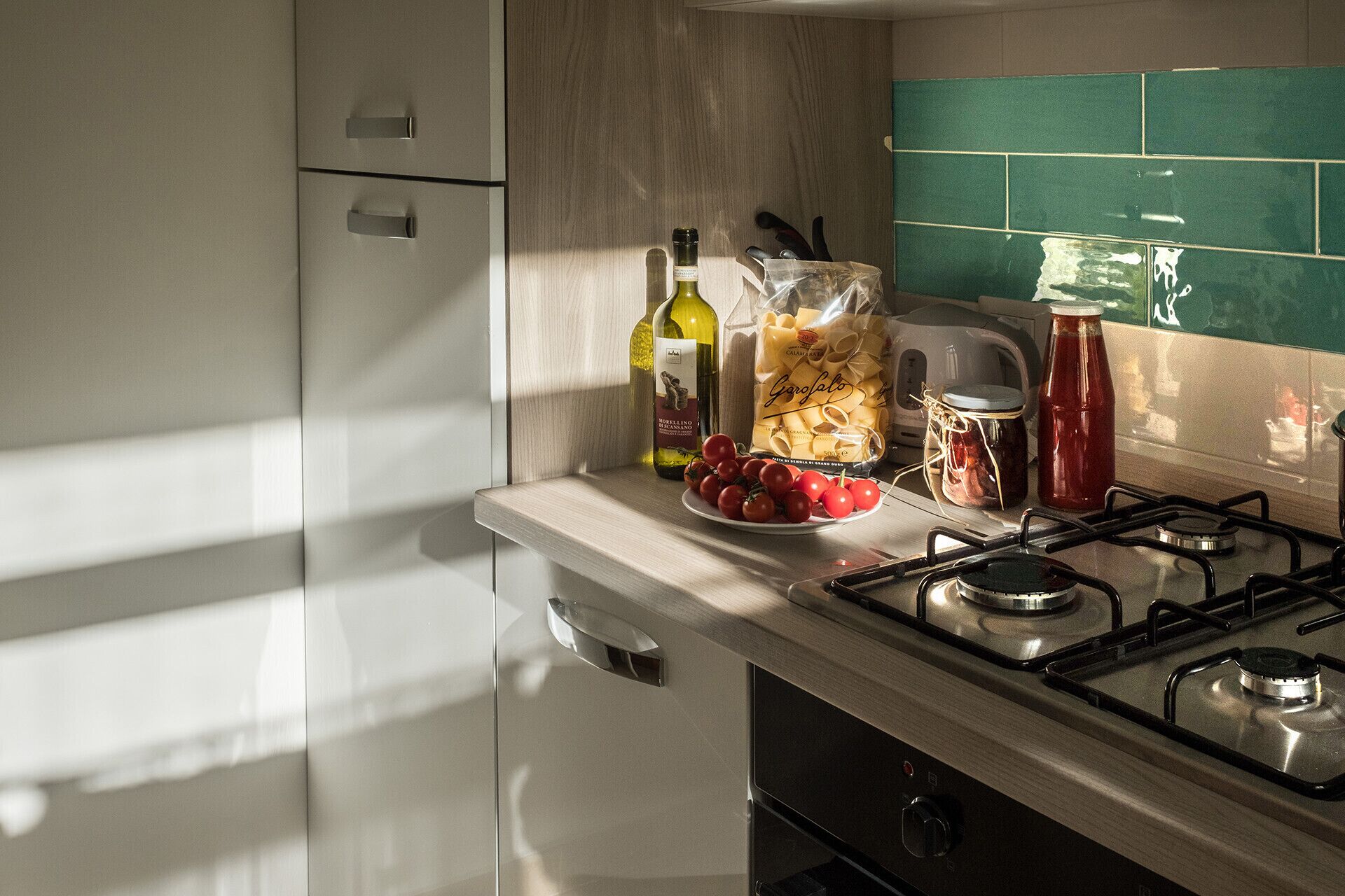 Een keuken met een gasfornuis, een fles olijfolie, een zak pasta, een pot saus en een bord met tomaten.