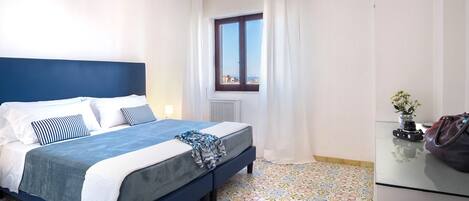 Une chambre d’hôtel avec un lit, une fenêtre avec des rideaux, un radiateur et un sol carrelé.