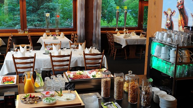 A restaurant with multiple tables set for dining, a buffet with various food items, and a view of greenery through the windows. - Fuxxbau Fischerbach (Fischerbach)