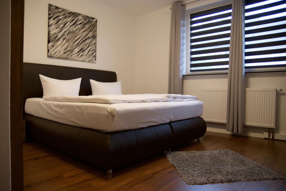 Un dormitorio con cama, cabecera, radiador, ventana con persianas y una alfombra en el suelo.