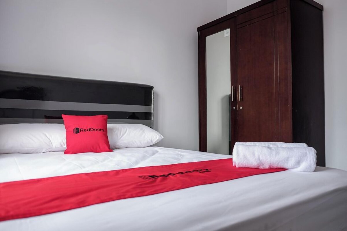 Ein Hotelzimmer mit einem Bett, einer roten Bettdecke, einem weißen Kopfkissen, einem roten Kissen, einem weißen Handtuch und einem Kleiderschrank.