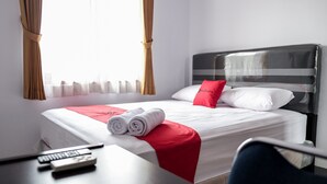 Una cama bien hecha con ropa de cama blanca y roja, una toalla enrollada al lado y un control remoto sobre una mesa.