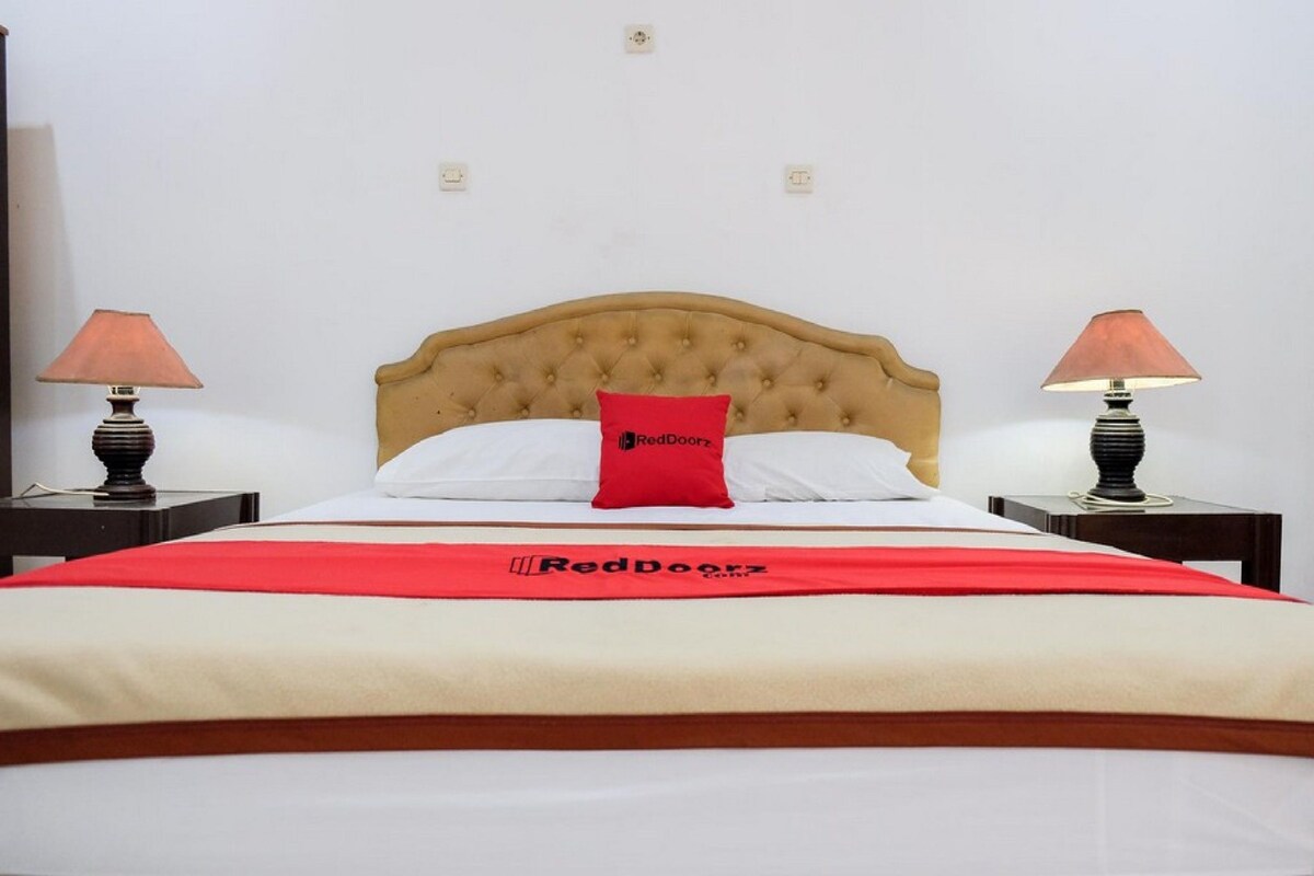 A bed with a tufted headboard, two bedside lamps, a red pillow, and a red blanket with a logo.