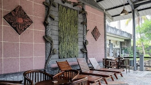 A covered outdoor seating area with wooden chairs and tables, a decorative wall with leaf patterns, and a tiled floor. - RedDoorz Plus near Jalan Baru Bogor (Bogor)