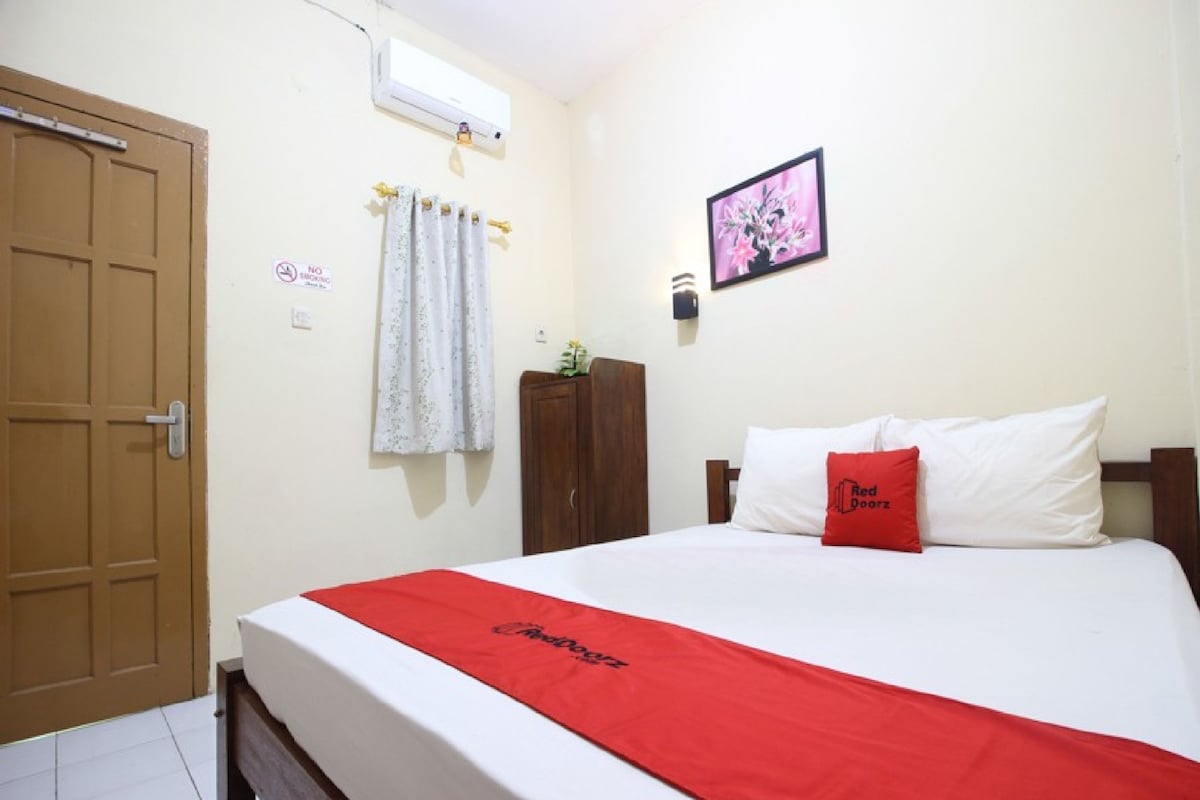 A hotel room with a bed, a wardrobe, a door, a wall-mounted air conditioner, and a picture of flowers.