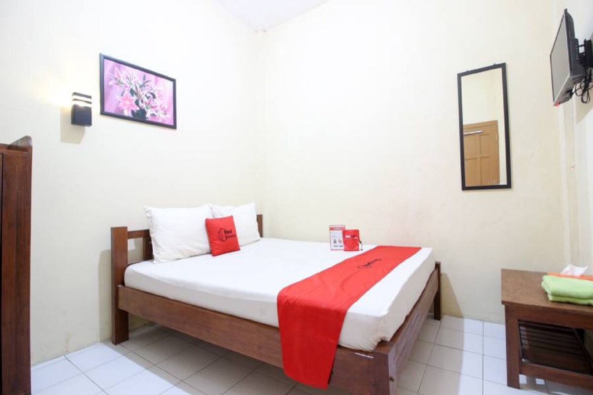 A single bed with white bedding and a red throw, a wooden bedside table with a green towel, a framed picture on the wall, and a mirror.