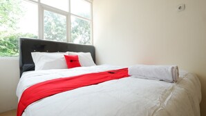 A neatly made bed with white bedding and a red accent, a window with a view of greenery, and a white wall with a power outlet. - RedDoorz near Akademi Kepolisian Semarang (Semarang)