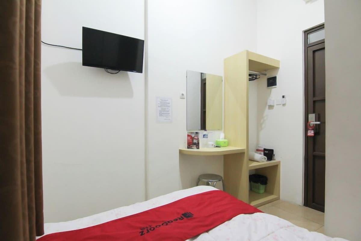 A hotel room with a bed, a TV mounted on the wall, a mirror, a small desk with a lamp, and a door.