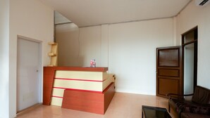 A reception desk with a wooden finish and a red accent, a brown leather sofa, and a glass coffee table in a room with white walls and a tiled floor. - RedDoorz near Akademi Kepolisian Semarang (Semarang)