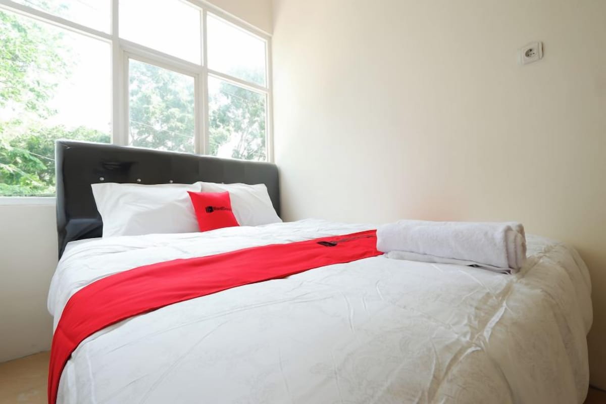 A neatly made bed with white bedding and a red accent, a window with a view of greenery, and a white wall with a power outlet.