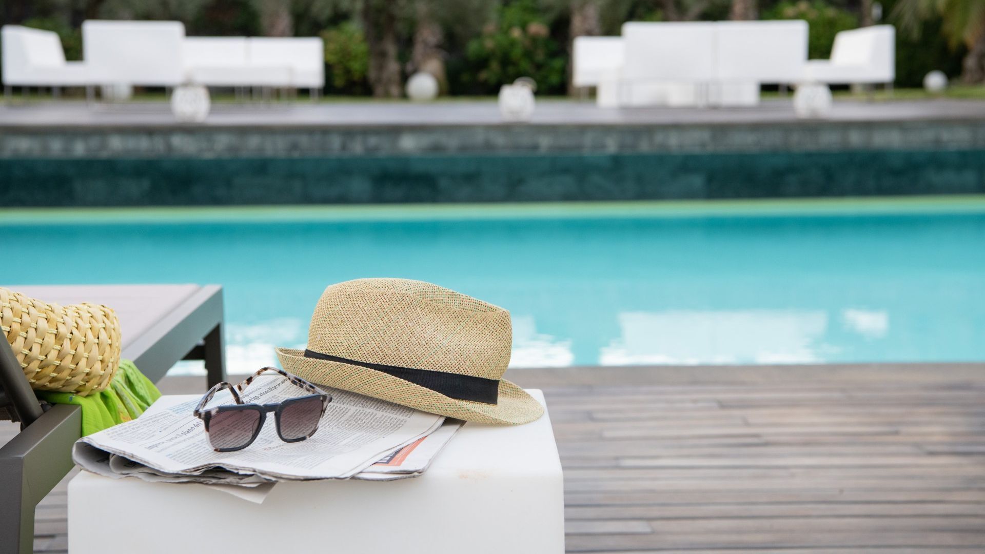 Zona de piscina con un sombrero, gafas de sol y un bolso tejido sobre una mesa.