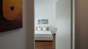 Een slaapkamer met een bed, wit beddengoed, een kleurrijk abstract schilderij aan de muur en een houten vloer.