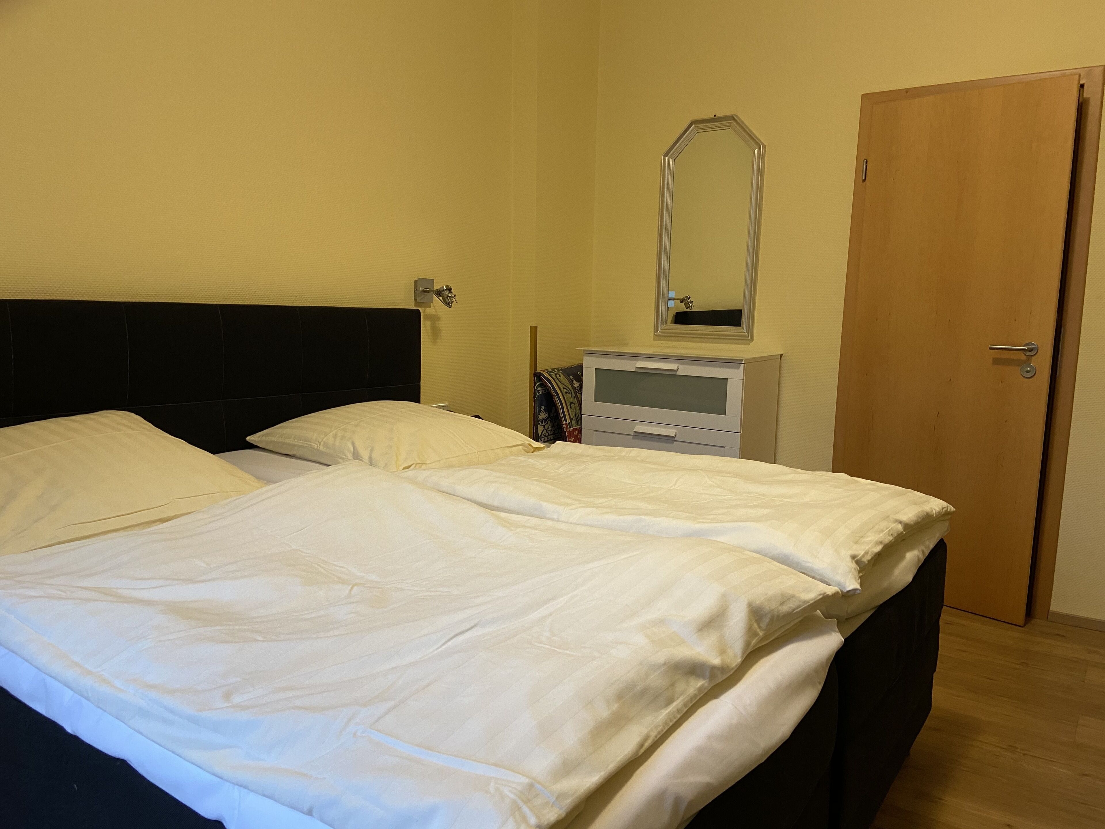 A double bed with white linens, a bedside table with a mirror, and a wooden door.