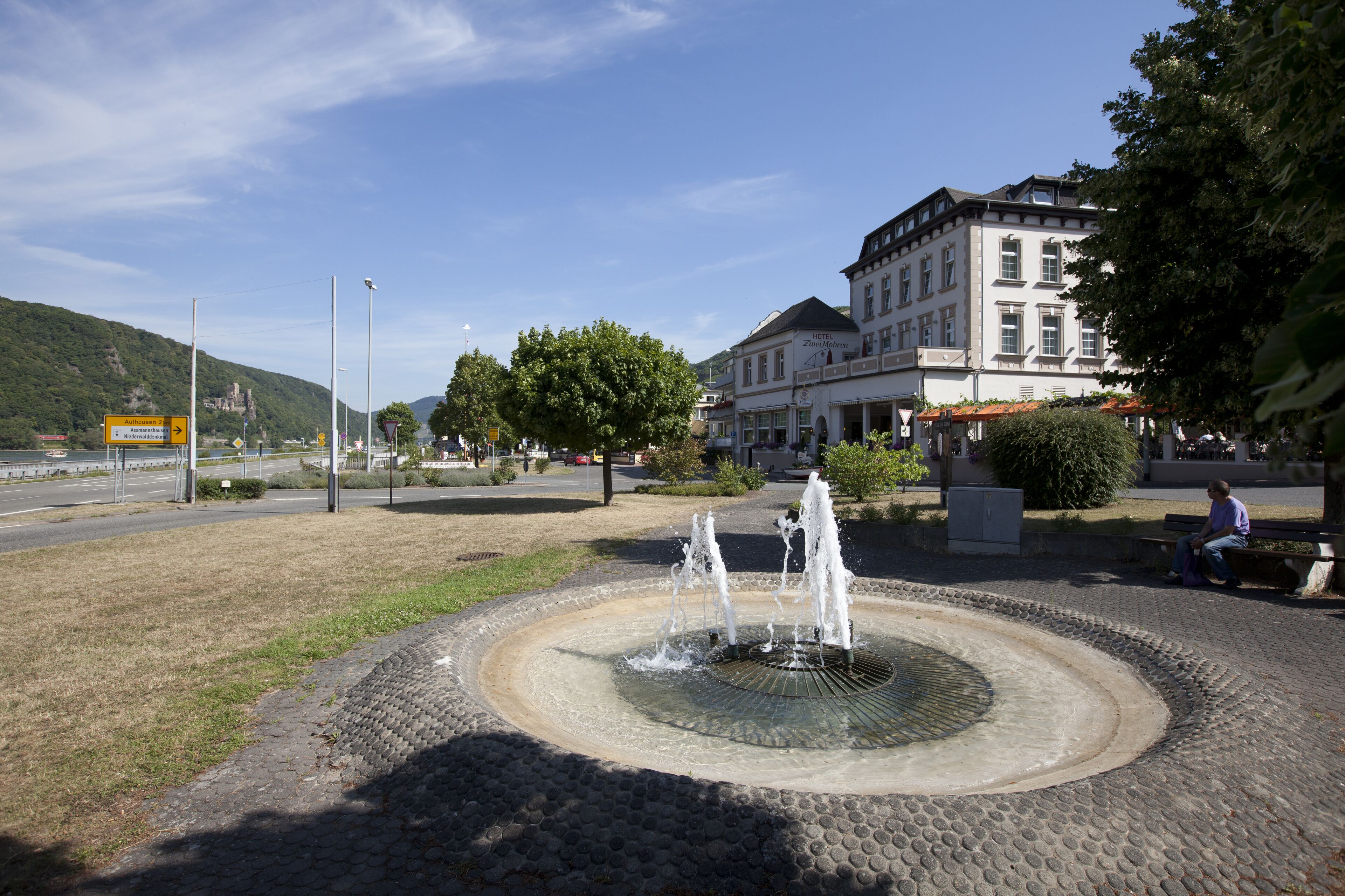 A public fountain with water jets in a circular basin, surrounded by cobblestone pavement. In the background, there is a multi-story building with a sign that reads "Hotel Zum Zorn."