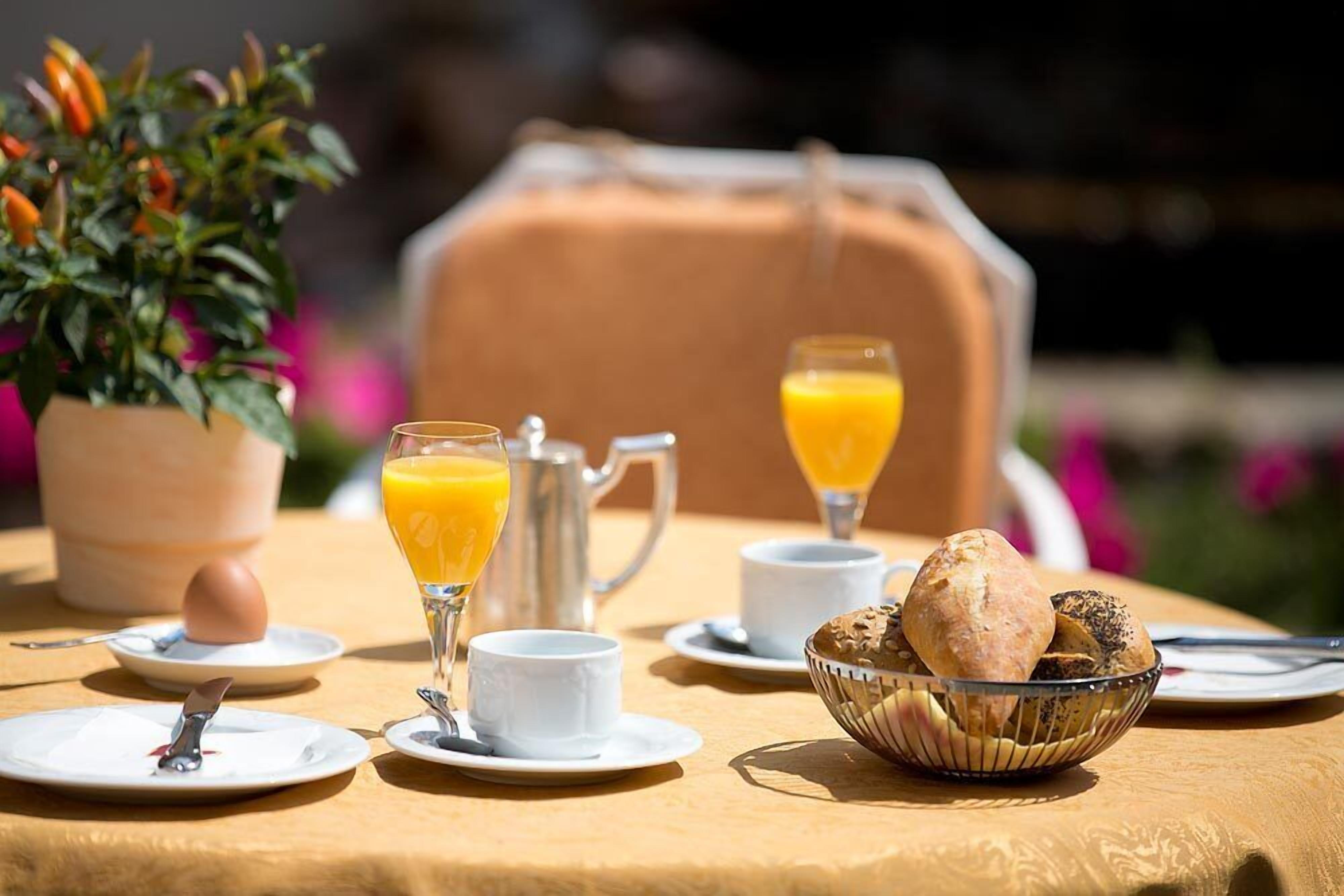 Une table mise pour le petit-déjeuner avec une carafe, deux verres de jus d’orange, un bol de pain et une plante en pot.