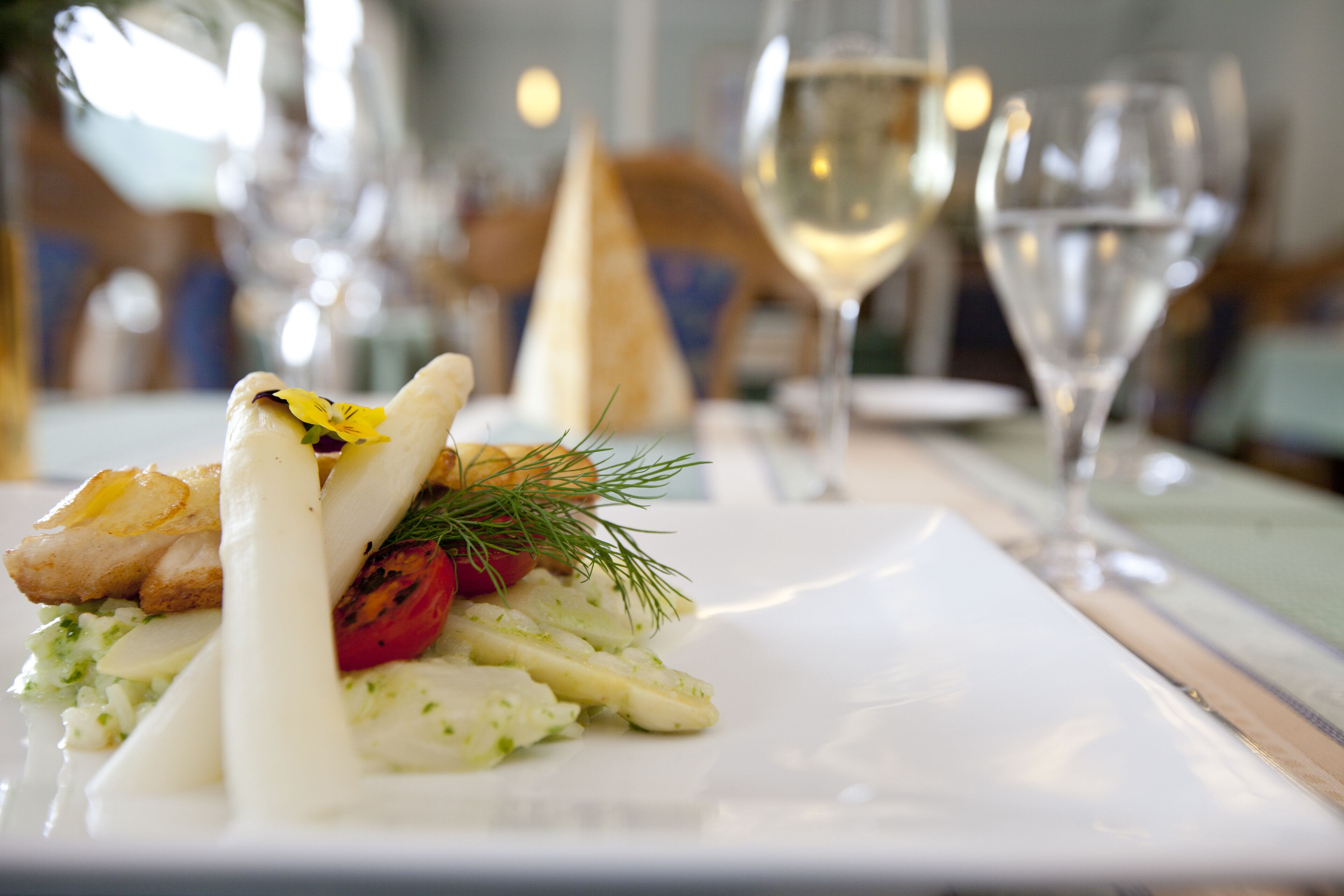 A dining table with a white plate of food, a glass of white wine, and a wooden chair.