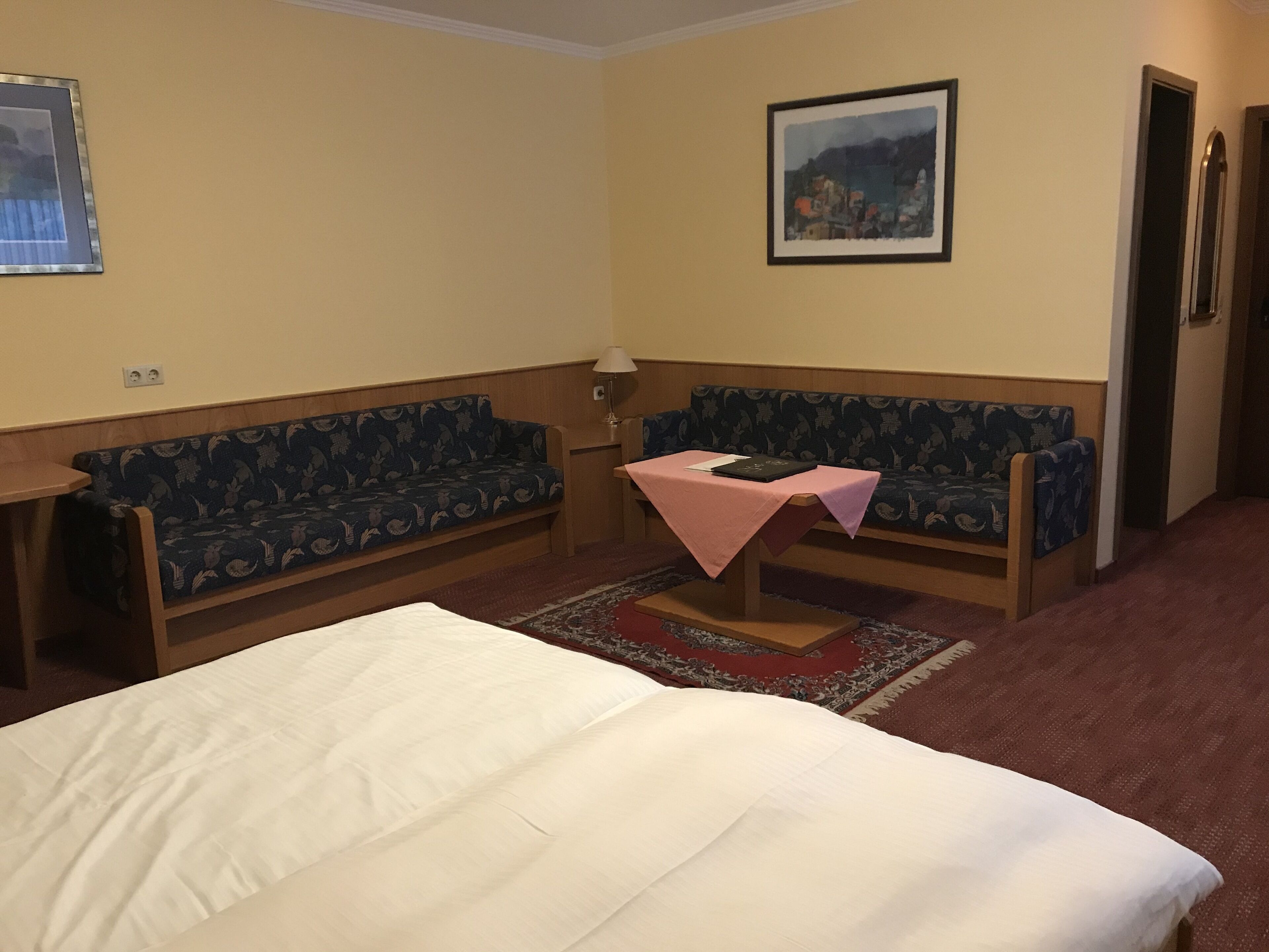 Habitación de hotel con cama, dos sofás, una mesita con mantel rosa, lámpara y un cuadro en la pared.