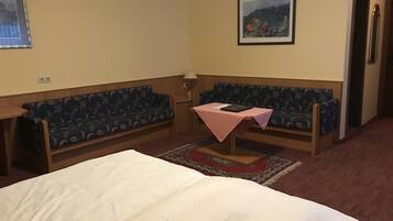 Habitación de hotel con cama, dos sofás, una mesita con mantel rosa, lámpara y un cuadro en la pared.