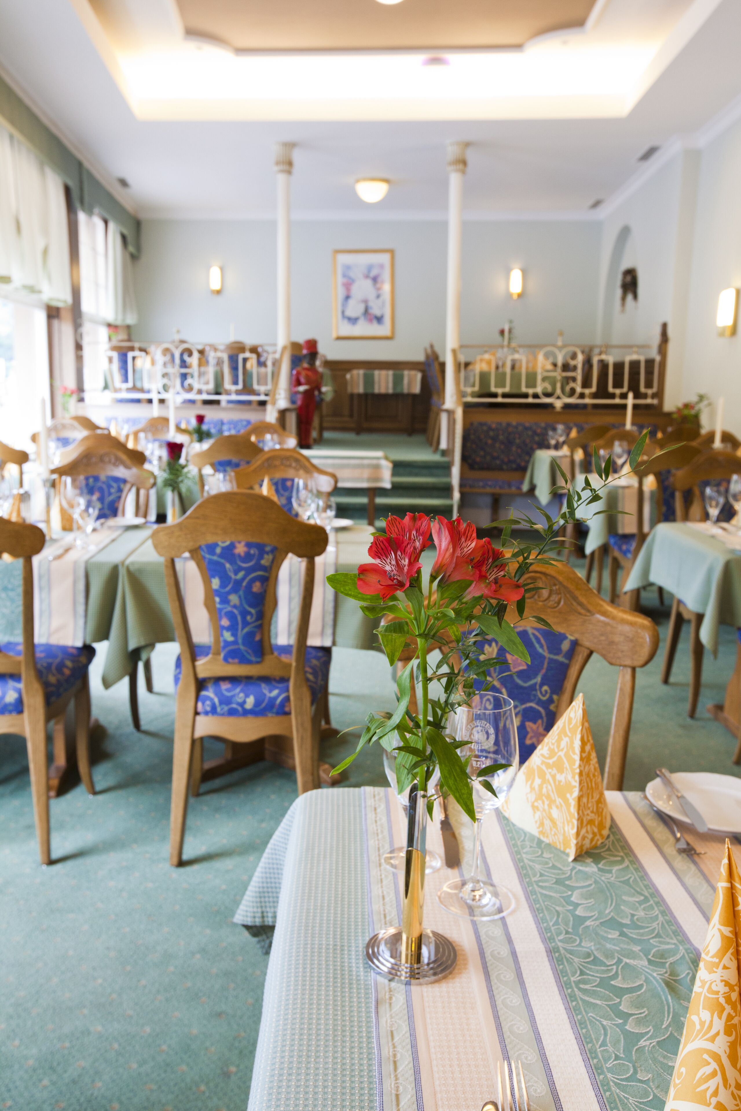 A restaurant with wooden chairs and tables, a vase with flowers, and a staircase in the background.