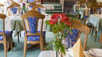A restaurant with wooden chairs and tables, a vase with flowers, and a staircase in the background.