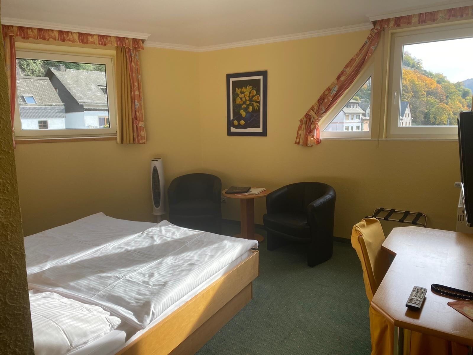 Ein Hotelzimmer mit Bett, Schreibtisch, Sessel, Fernseher und einem Fenster mit Blick auf Gebäude und Bäume.