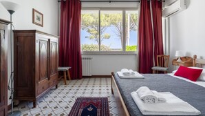 Een hotelkamer met een bed, een houten kledingkast, een radiator, een raam met gordijnen en uitzicht op bomen.