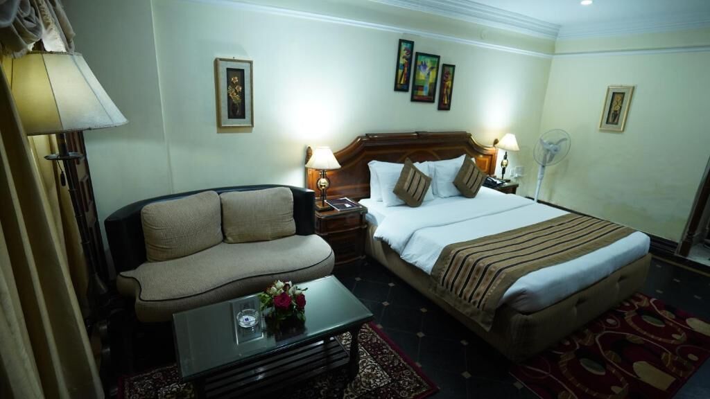 A hotel room with a bed, a sofa, a table with a vase of flowers, and two wall-mounted lamps.
