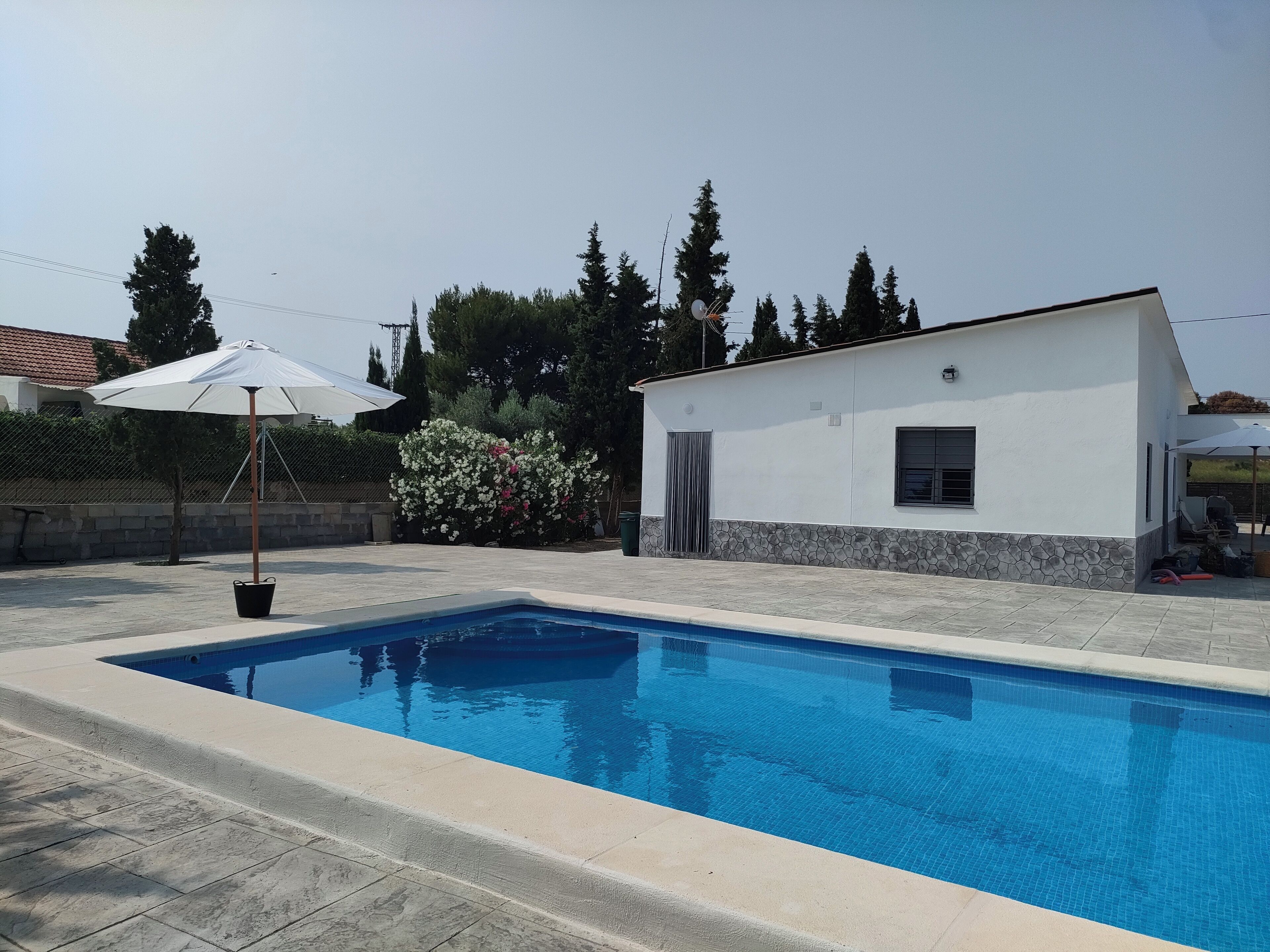 Une piscine bordée de pierres, un bâtiment blanc avec une fenêtre, et une clôture avec des arbres en arrière-plan.