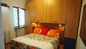 Un dormitorio con un armario de madera, una cama con colcha estampada, dos lámparas de noche y una ventana con cortinas.