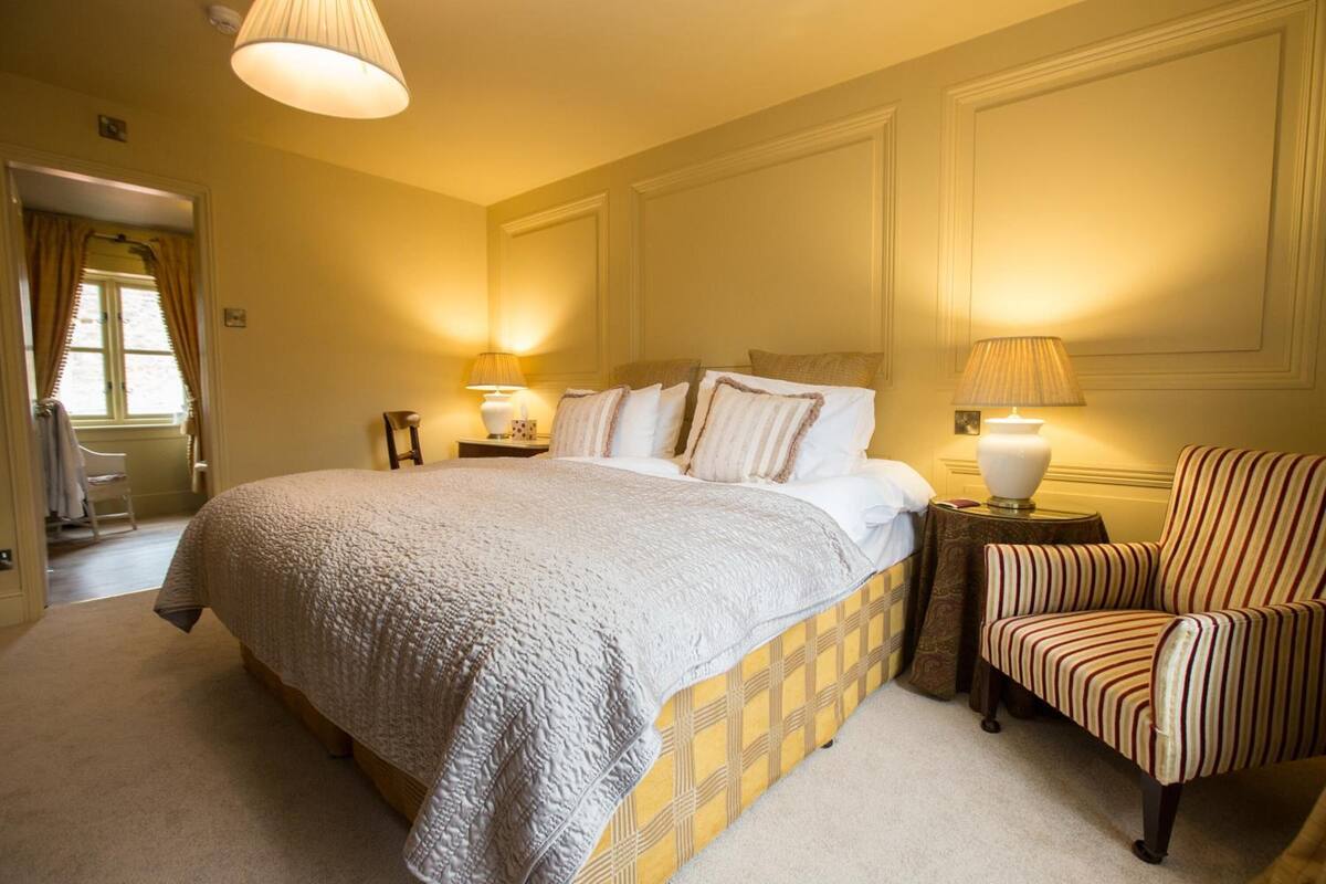 A hotel bedroom with a large bed, two bedside tables with lamps, a striped armchair, and a door leading to another room.