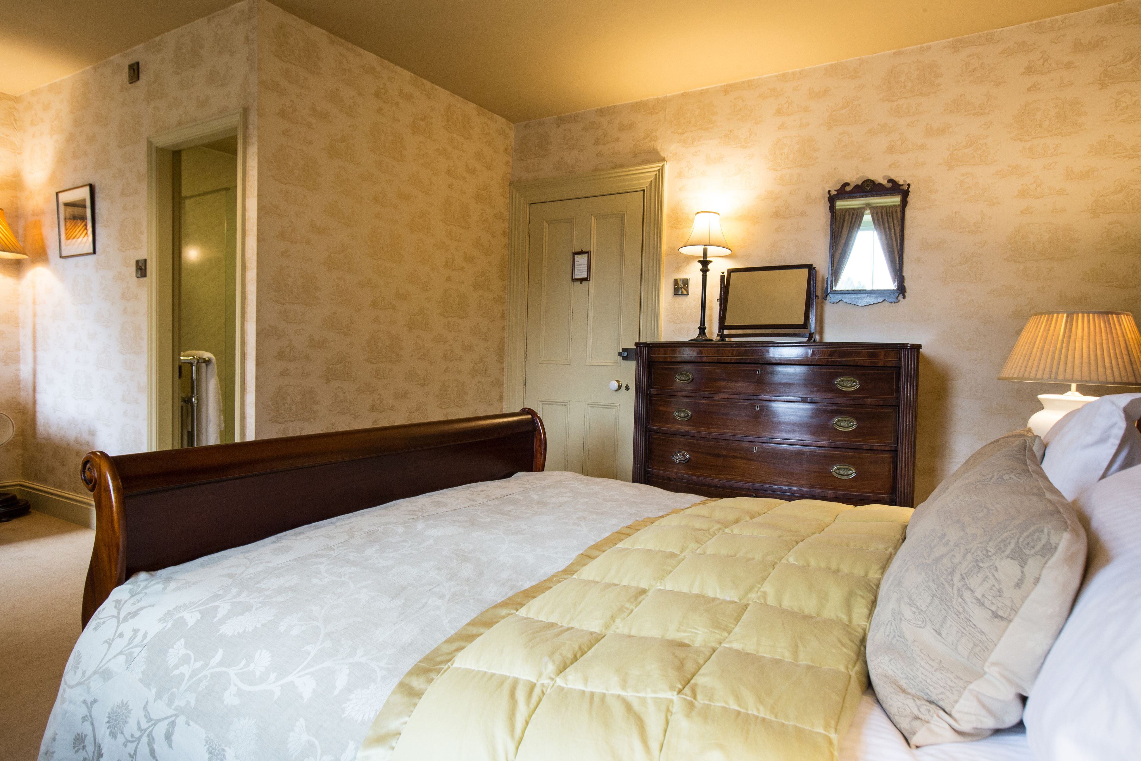 A neatly made bed with a quilted cover, a wooden dresser with a mirror, and a door leading to another room.