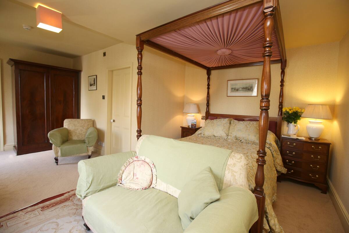A four-poster bed with a canopy, a nightstand with lamps, a chair, and a door in a bedroom.