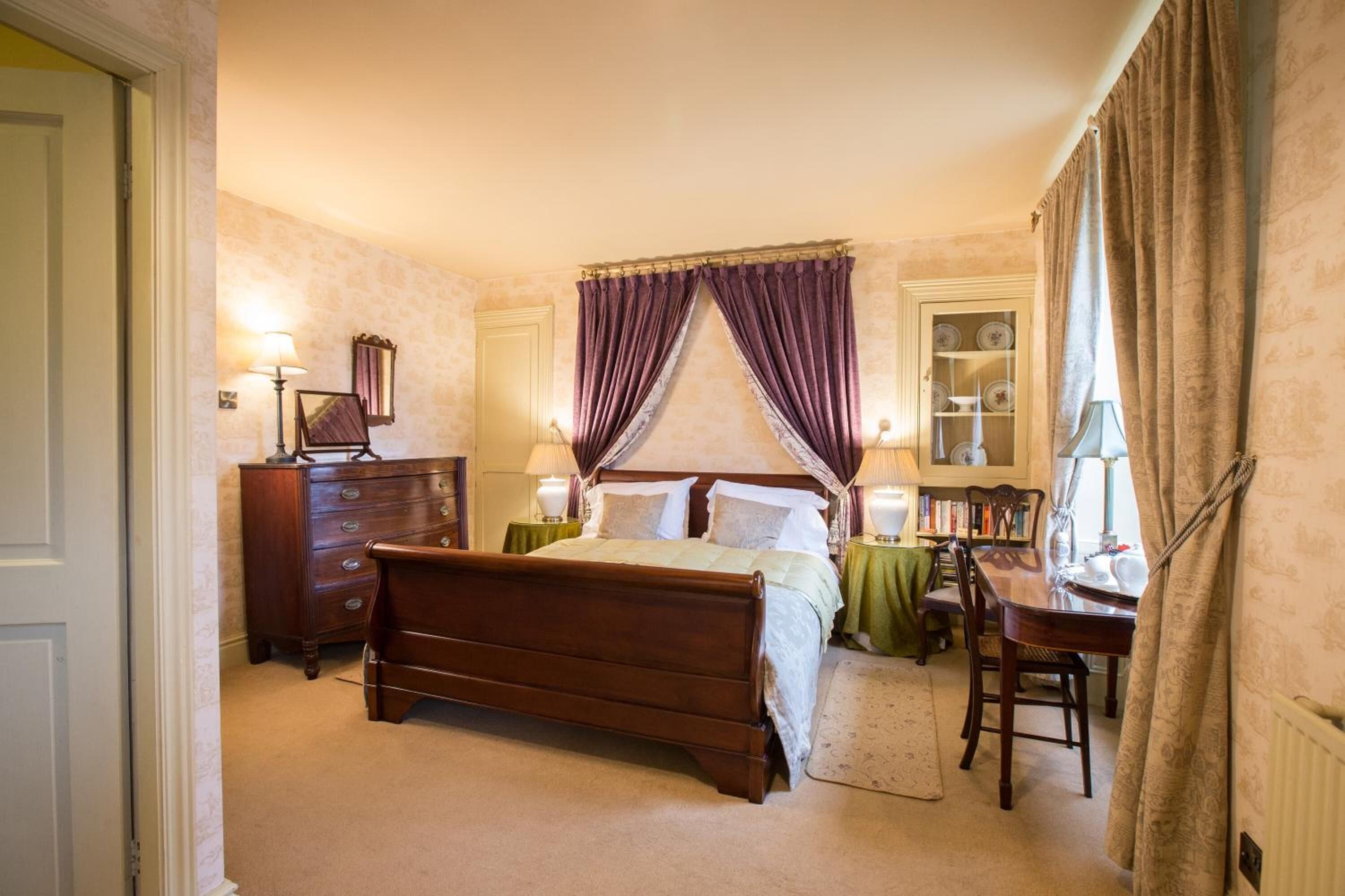 A bedroom with a wooden bed, a dresser, a desk, and a chair. There is a window with curtains and a bookshelf with books.