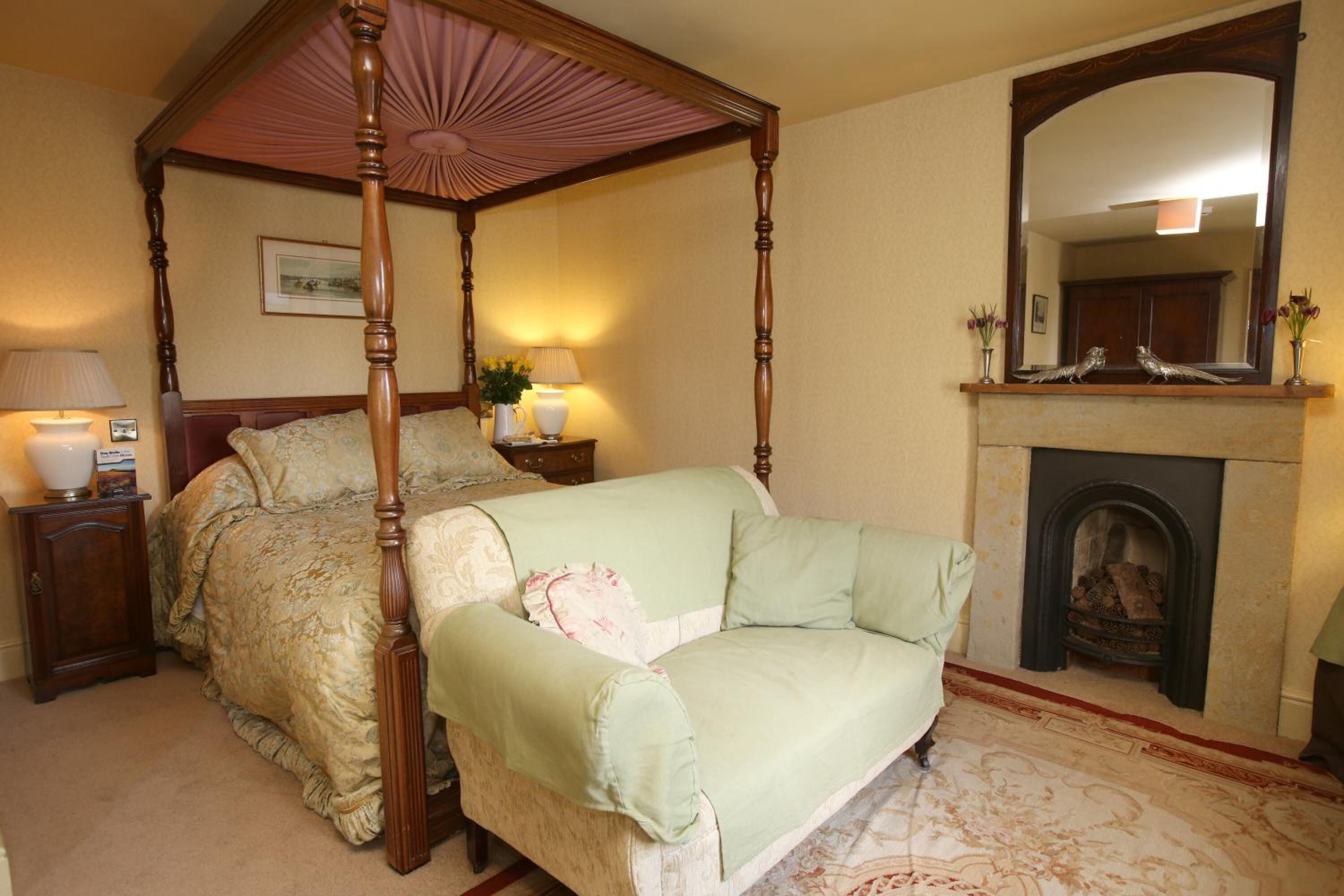 A four-poster canopy bed with a green upholstered sofa, a bedside table with lamps, a fireplace, and a mirror above it.