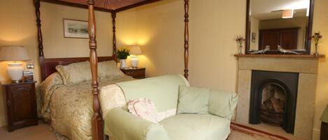 A four-poster canopy bed with a green upholstered sofa, a bedside table with lamps, a fireplace, and a mirror above it.