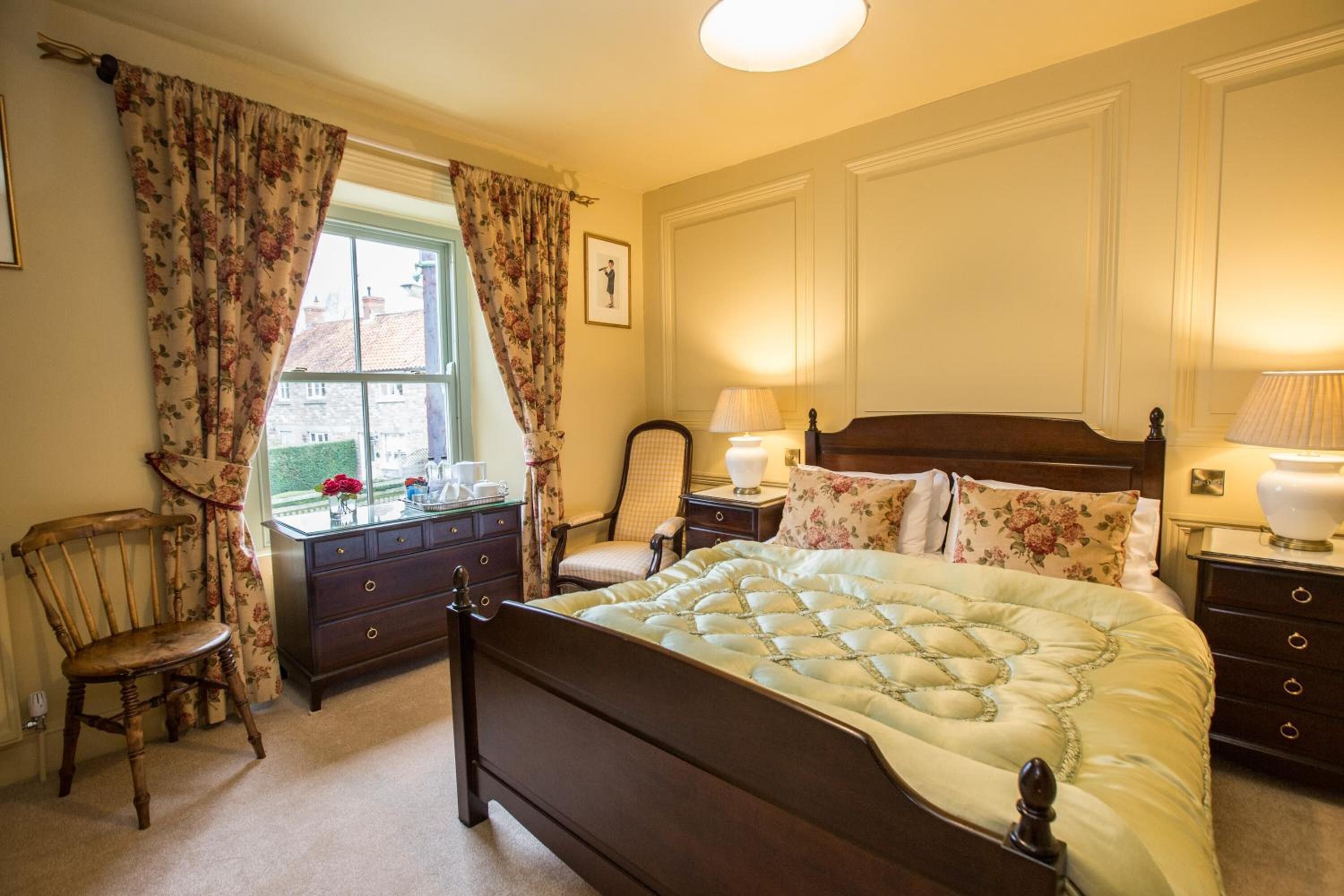 A bedroom with a large bed, a wooden nightstand, a chair, a dresser, and a window with curtains.