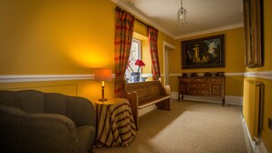 A room with a sofa, a small table with a lamp, a bench, a window with curtains, a cabinet, and a painting on the wall. - The Cornmill (York)