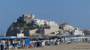 Una ciudad costera con una estructura imponente al estilo de castillo en una colina, sombrillas y sillas de playa.