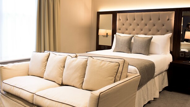 A hotel room with a bed, a sofa, a nightstand, and a window with curtains. - The County Aparthotel (Newcastle-upon-Tyne)